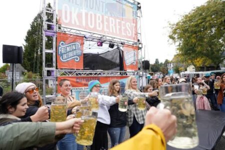 People stand outdoors at Oktoberfest, holding large mugs of beer in a group toast. A stage with “Fremont Oktoberfest” banners is in the background. Trees and festival booths are visible on a cloudy day.