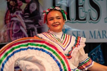 A woman in traditional Mexican dress dances, holding a colorful skirt with rainbow stripes, smiling during a performance on stage.