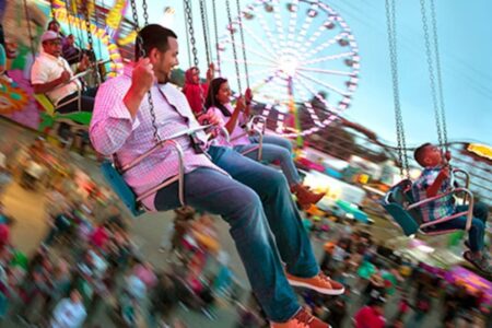 People of various ages enjoying a colorful carnival swing ride, with a ferris wheel and a crowd in the background, creating a sense of motion and excitement.