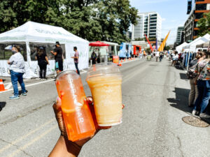 A hand holds two bubble tea drinks at an outdoor street market, with white vendor tents, people, and buildings visible in the background under a sunny sky.