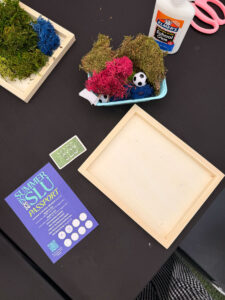 A tabletop displays moss, glue, a soccer ball, wooden frames, a “Made in SLU” pin, and a “Summer in SLU Passport” flyer with event information and a QR code. Craft supplies are arranged for an activity.