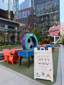 Outdoor seating area with orange chairs, foosball table, and a large soccer ball sculpture. A sign promotes “Made in SLU” event. In the background, buildings and the pink Elephant Car Wash sign are visible.