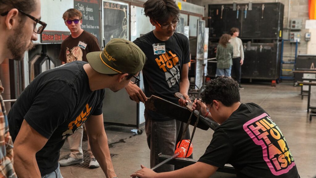 Three people wearing “Hill Top ISD” shirts work together shaping molten glass in a workshop, while others watch in the background.