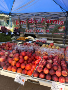 A farmers market stall displays peaches, apples, and grapes in bags and loose. Amador Farms banner hangs above the stand under a blue tent. Prices are visible on signs.