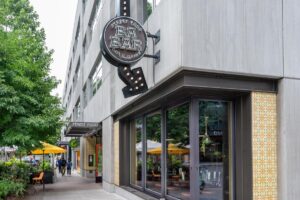A corner view of Ba Bar restaurant with outdoor seating, people walking by, and yellow umbrellas on the patio. The sign reads “Ba Bar Street Food, Cocktails, Dining.”.