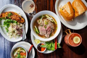 A table with pho soup, banh mi sandwiches, fried fish with slaw, a dipping sauce, a cocktail with star anise, and a copper cup with a lemon slice, all arranged on a dark wooden surface.