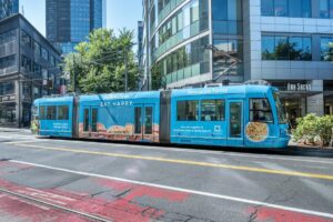 A blue streetcar with Mendocino Farms ads reading “EAT HAPPY” travels through a city street with modern glass buildings and trees on a sunny day.
