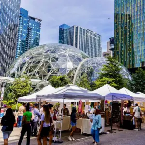 People browse outdoor market stalls with white tents in front of large glass domes and tall modern buildings on a sunny day. Trees and greenery are visible among the urban architecture.