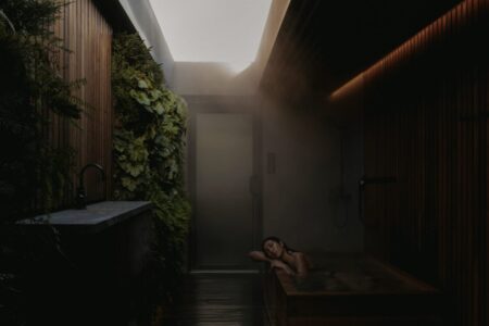 A woman relaxes in a wooden bathtub in a dim, steamy spa room with wooden walls, a vertical garden, a sink, and a skylight above letting in natural light.