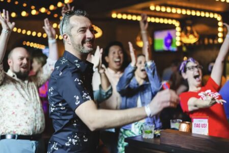 A man in a floral shirt throws a dart while a cheering crowd watches in a lively indoor setting with string lights and drinks on the table.