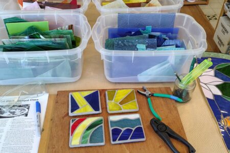 Four colorful stained glass coasters are on a table with pliers, glass cutter, a pen, a class paper, and bins of colored glass sheets. A partially finished stained glass piece is at the right edge.