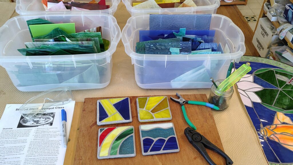 Four colorful stained glass coasters, pliers, a jar with tools, and bins filled with glass pieces are on a worktable. An instructional paper and a pen are on the left side of the table.