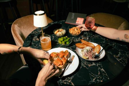 Two people share a meal at a black marble table with drinks, appetizers, olives, and toasted bread with toppings under warm lighting. One person reaches toward the food.