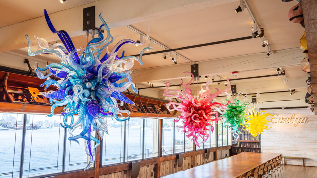 Colorful glass chandeliers resembling sea creatures hang from a ceiling in a waterfront dining area with large windows and a long wooden table.