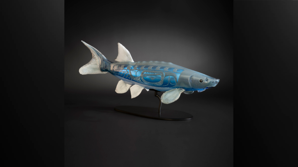 A realistic fish sculpture with blue Indigenous designs on its body is mounted on a black stand, set against a dark gradient background.