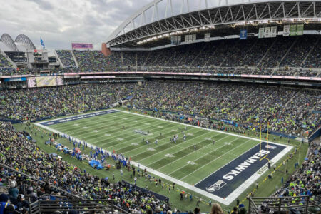 A packed football stadium with fans watching a game between the Seattle Seahawks and another team. The field is marked with Seahawks logos, and the retractable roof is partially visible under a cloudy sky.