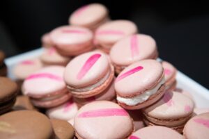 A pile of pink macarons with white filling and pink stripes on top, stacked next to a few brown macarons, displayed on a white plate.