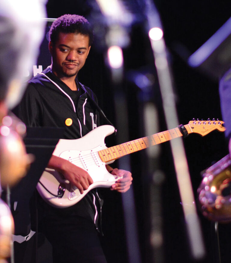 A musician wearing a black shirt plays a white electric guitar on stage, looking to the side. Other instruments and musicians are slightly visible in the foreground.