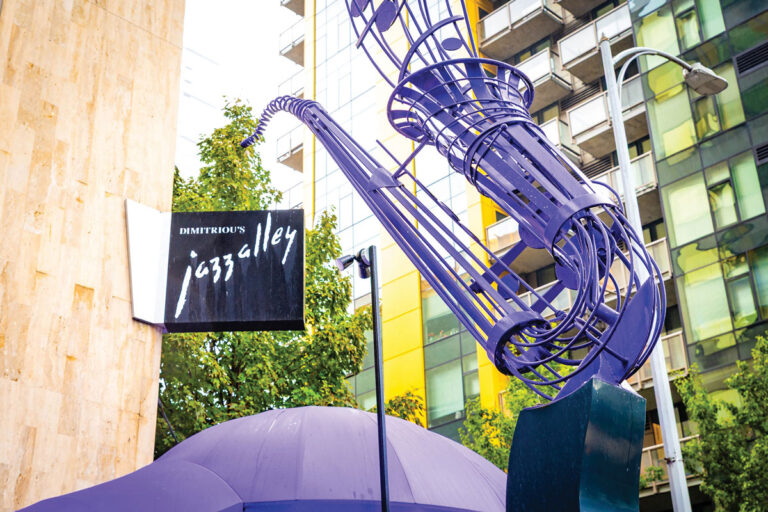 A large purple saxophone sculpture stands next to the Dimitriou’s Jazz Alley sign, with tall modern buildings and trees in the background.