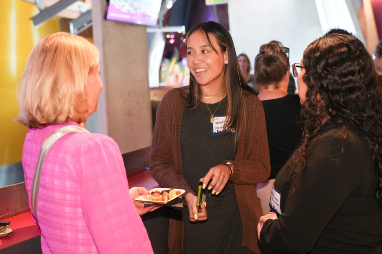 Three women in a conversational group at a Become a Visit Seattle Partner event, with the woman in the center wearing a name tag and holding a small plate of food.