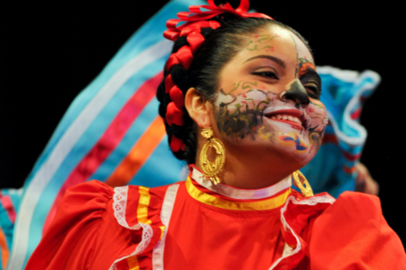 A dancer in a red dress with a blue ruffled skirt performs with her face painted in a traditional Day of the Dead style.