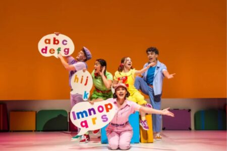 Five performers in colorful outfits on stage hold speech bubble cutouts featuring groups of letters from the alphabet.