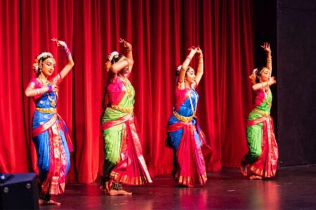 Four dancers in colorful traditional attire perform on a stage in front of red curtains.