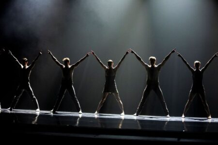 Five dancers in silhouette stand in a line on a stage with arms raised in a V-shape.