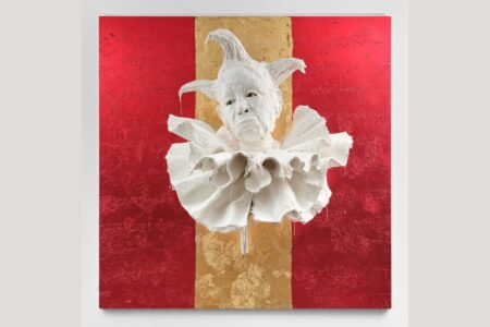 A white sculpture of a jester head with a ruffled collar mounted on a square panel divided into red side sections and a gold center stripe.