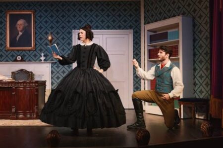 A woman in a period-style black gown holds a book while a man in a vest kneels before her on a stage set.