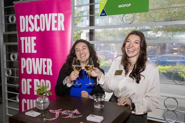 Two women smile and toast with glasses of white wine in front of a pink “Discover the Power of Pop Culture” sign, representing those who become a Visit Seattle partner.
