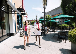 A woman and a man walk along an outdoor shopping mall sidewalk past storefronts and café tables with umbrellas.